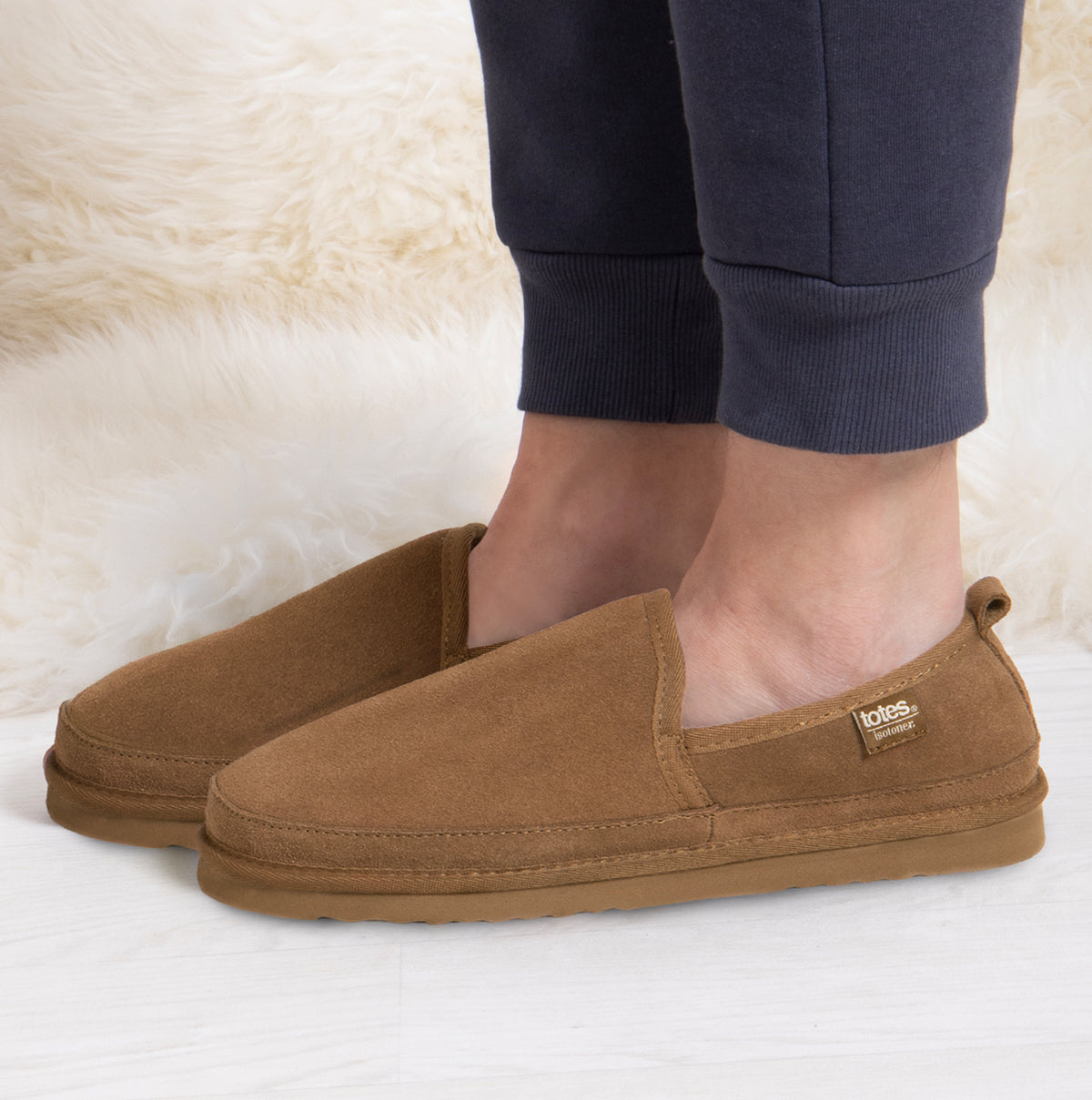 Totes Isotoner REAL SUEDE FULL BACK Mens Full Back Slippers Tan side view on a model's feet with casual pants.